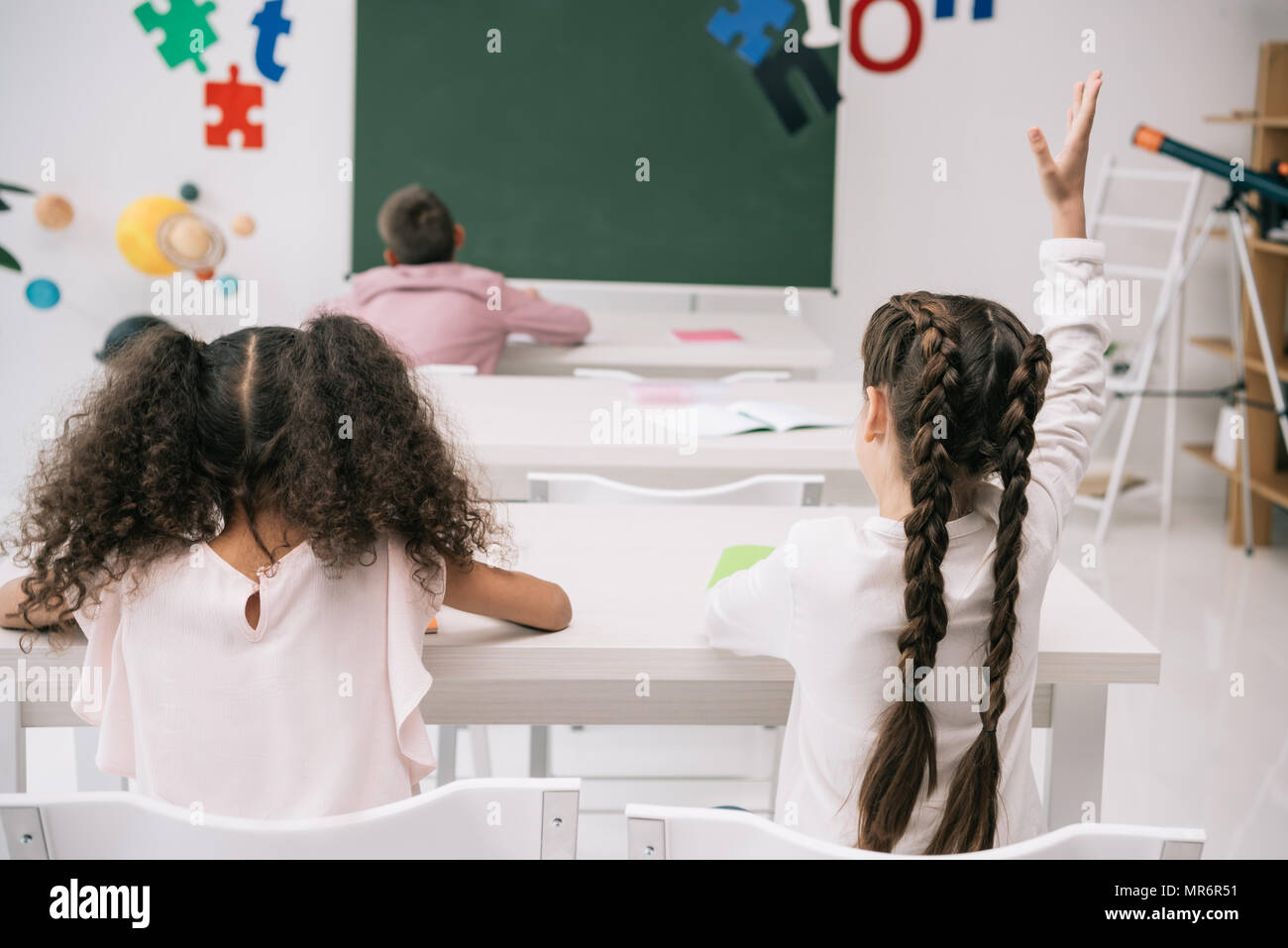 Back view of pupils sitting at desks and schoolgirl raising hand at ...