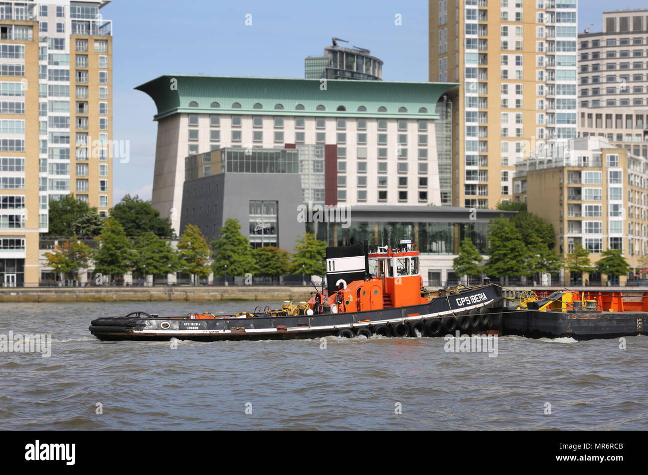 A tug boat pushed barges containing refuse containers along the River Thames in London, UK, passing the Canary Wharf Development. High tide, sunny day. Stock Photo