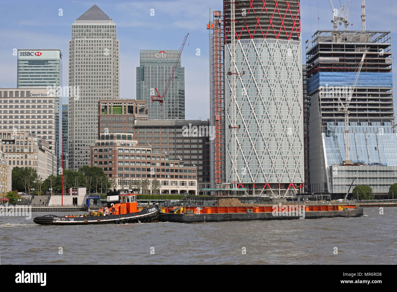 A tug boat pushed barges containing refuse containers along the River Thames in London, UK, passing the Canary Wharf Development. High tide, sunny day. Stock Photo