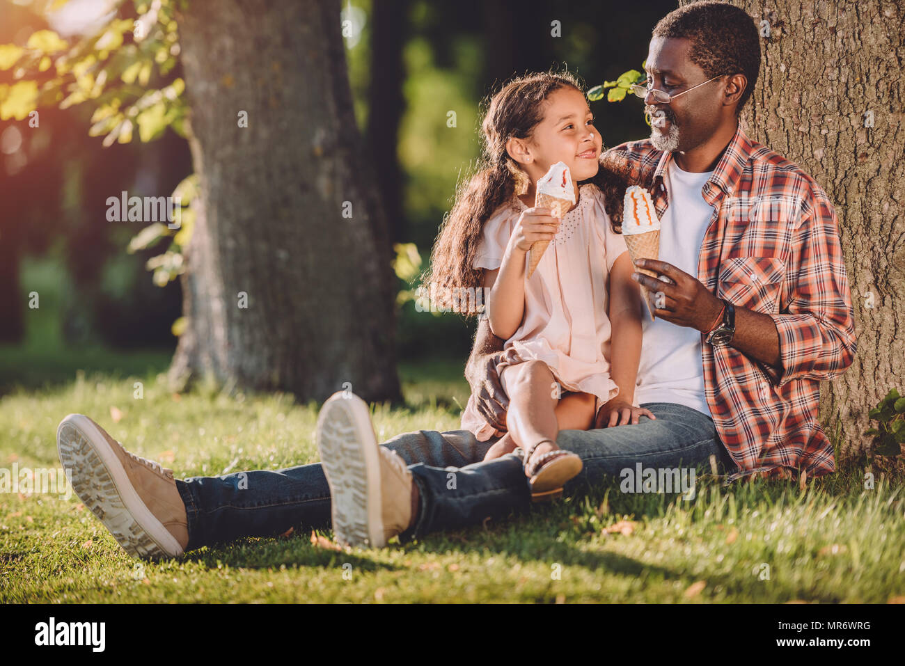 happy african american granddaughter and grandfather eating ice cream in cones while sitting on grass in park Stock Photo