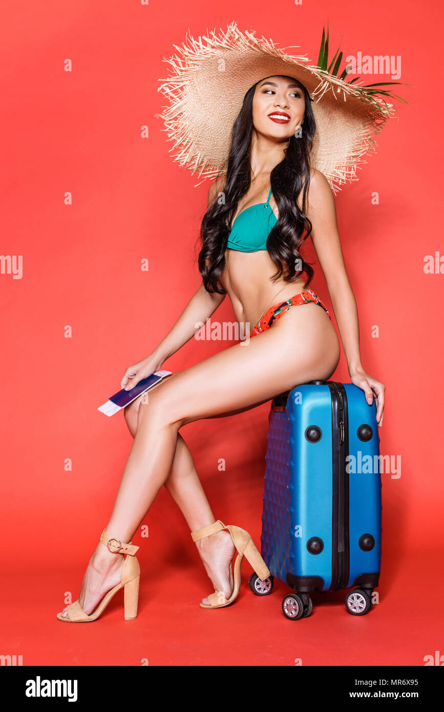 Full-length shot of beautiful asian woman in bright swimsuit and beach hat sitting on a suitcase and holding tickets Stock Photo