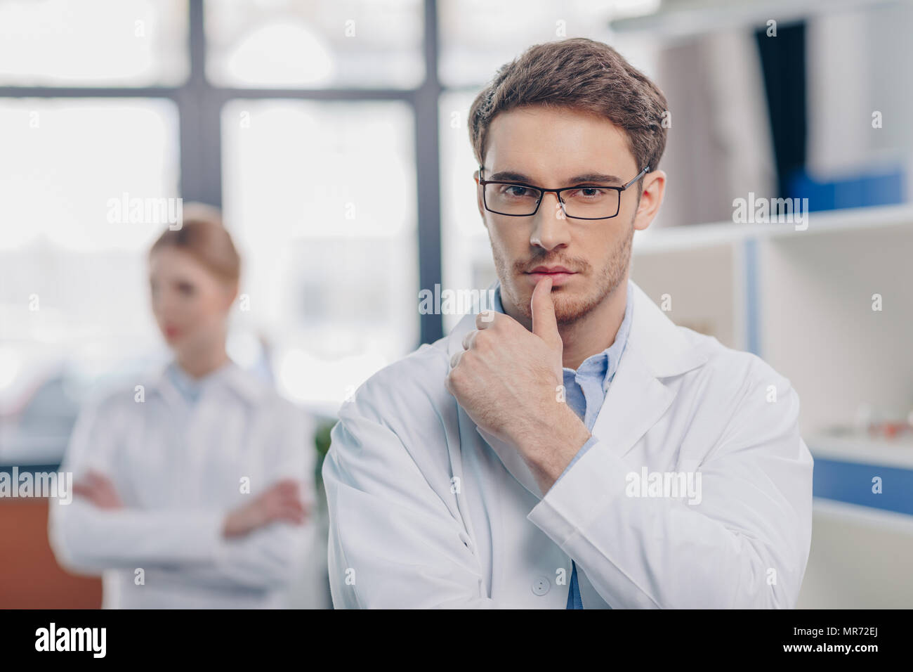 male pensive chemist in white coat in laboratory with colleague behind