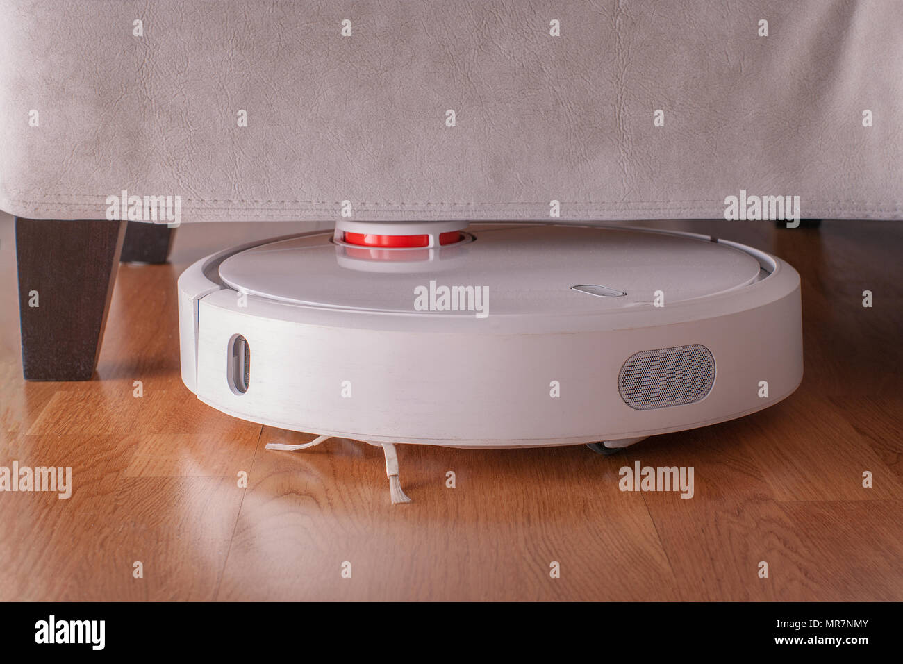 Robotic vacuum cleaner runs under sofa in room on laminate floor. Robot