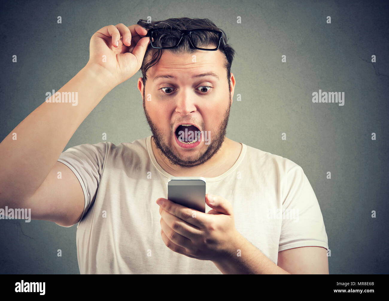 Super amazed young man with overweight reading shocking news on smartphone holding glasses up with mouth opened. Stock Photo