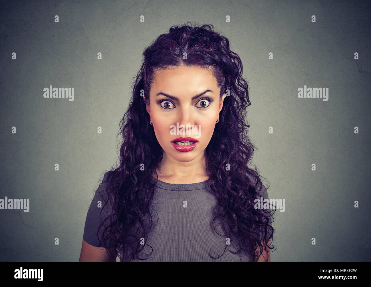 Young stunned brunette with curls holding eyes wide opened and looking scared at camera on gray. Stock Photo