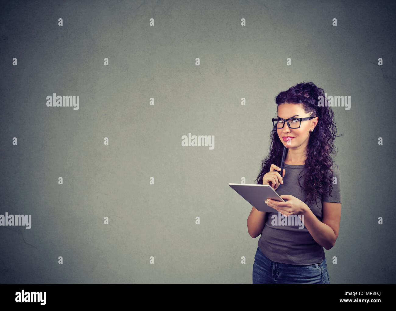 Stylish brunette thinking happily of new ideas while holding pen and using tablet on gray backdrop. Stock Photo