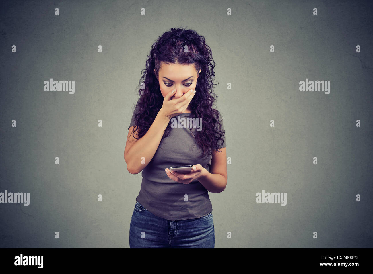 Young amazed girl covering mouth and reading news on smartphone having strong reaction on gray background. Stock Photo