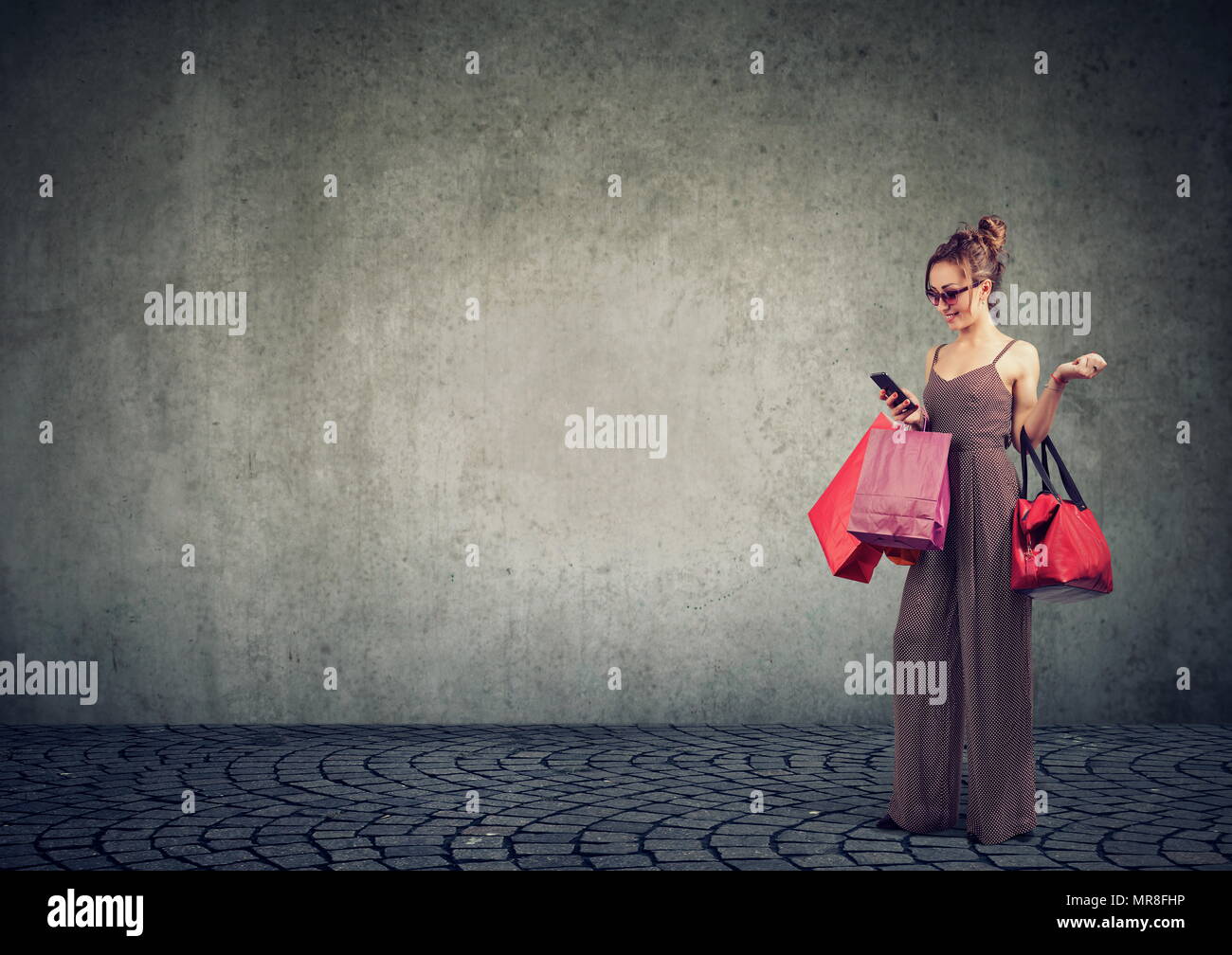 Excited trendy girl with shopping bags using smartphone checking prices online in the Internet on smartphone smiling on gray background. Stock Photo