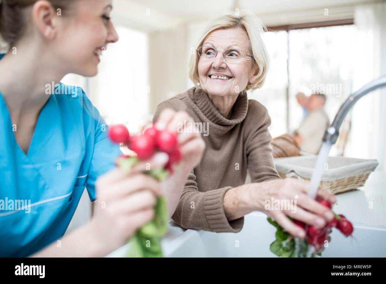Female care worker washing radishes with senior woman. Stock Photo