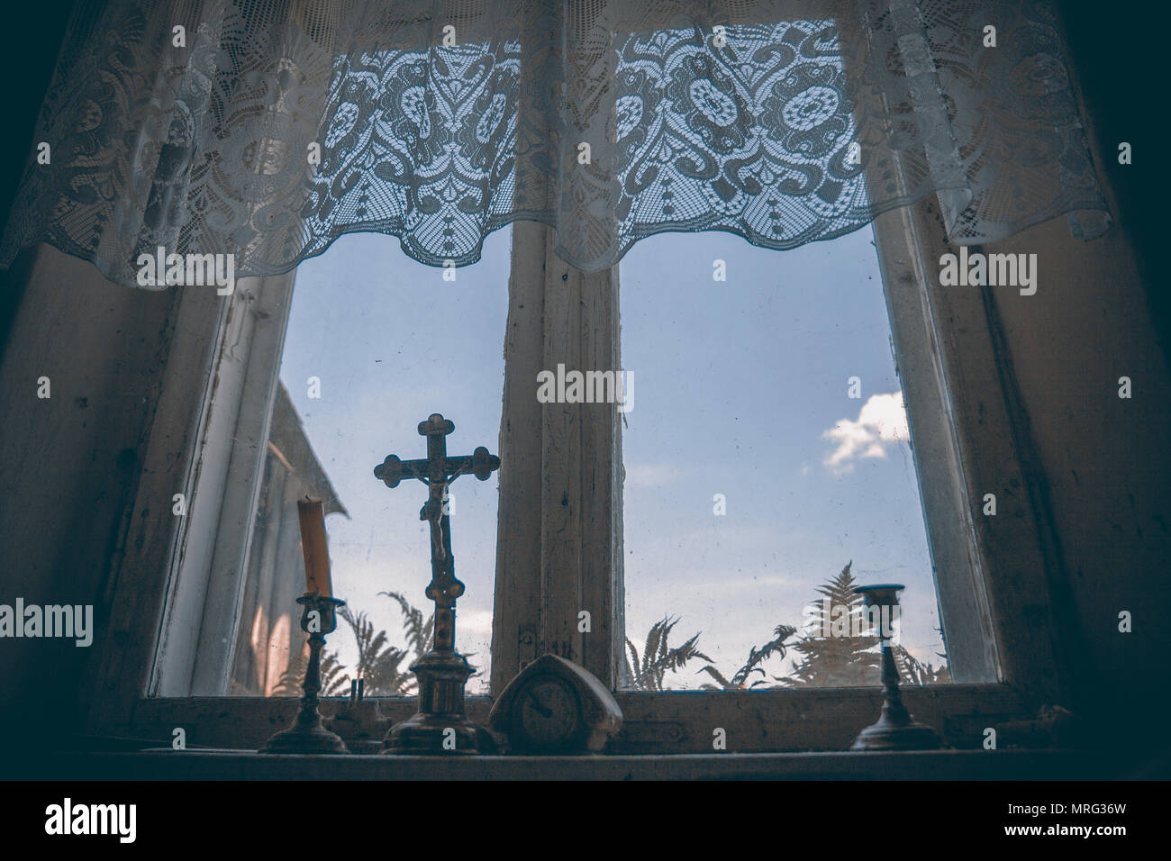 Candlesticks and crucifix standing under net curtain on an old oooden window. Stock Photo
