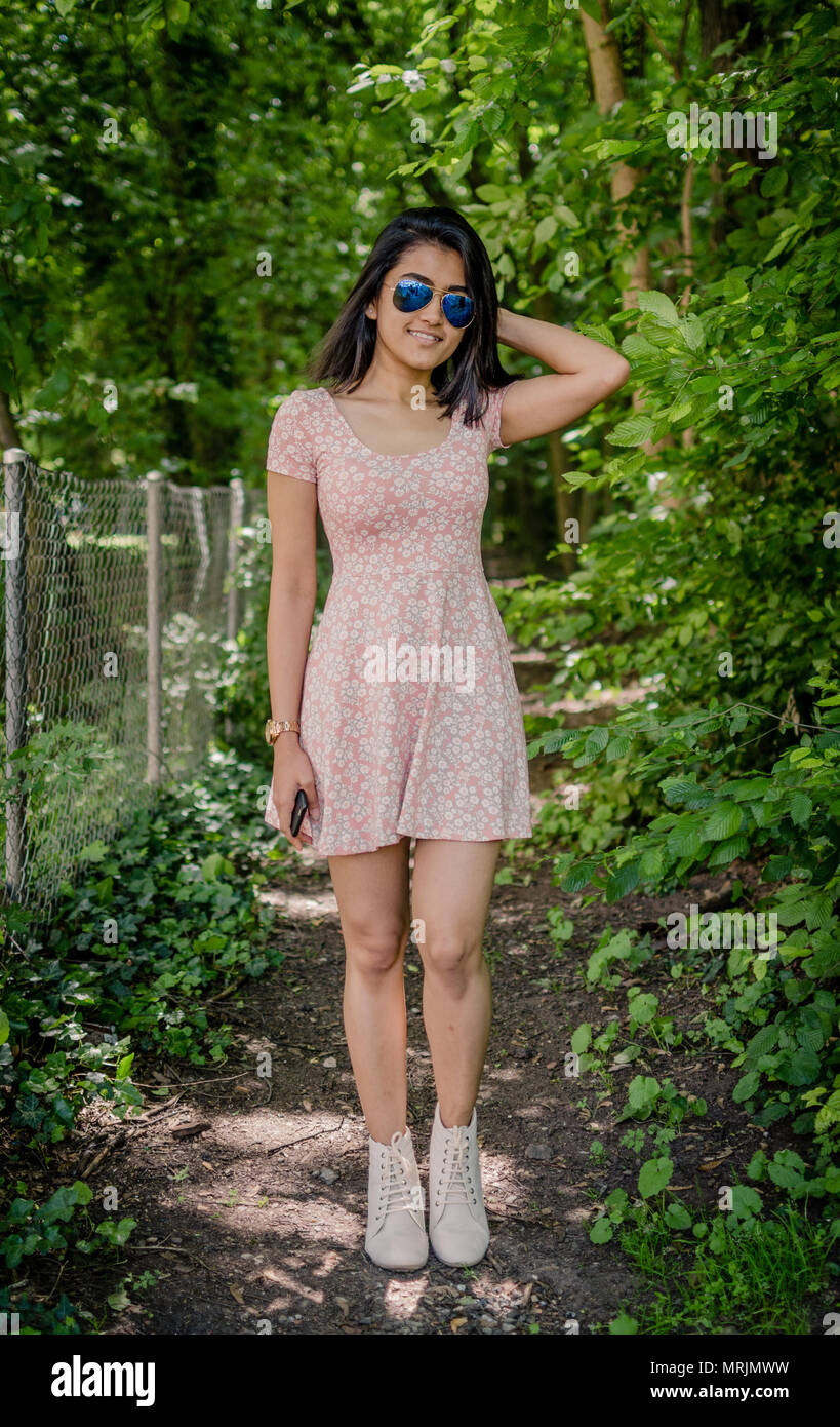 Full length portrait of a cute smiling girl in pink one piece dress, wearing blue aviator sunglasses, posing on a forest path / nature background. Stock Photo