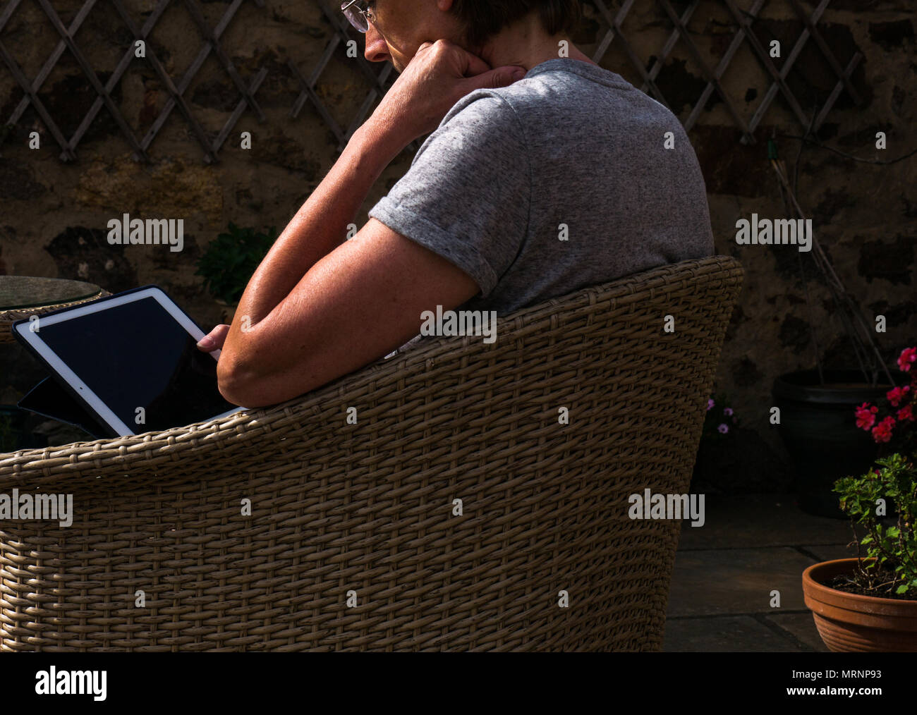 Side view profile of suntanned mature woman with reading glasses sitting in wicker garden chair on patio with flower pots looking at iPad screen Stock Photo