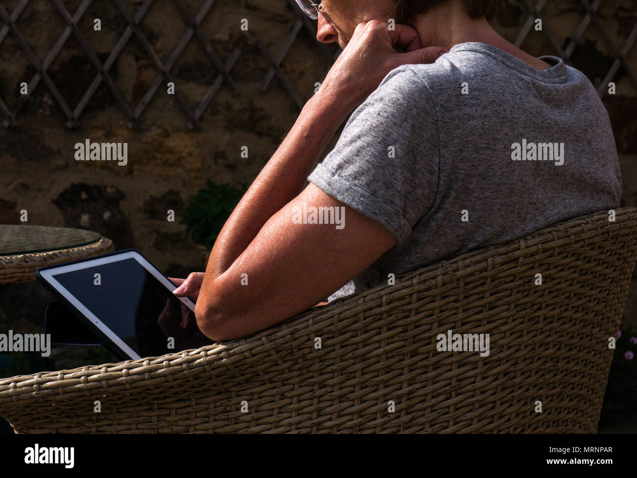 Sunlit side view profile of suntanned mature woman with reading glasses sitting in wicker garden chair on patio looking at iPad screen Stock Photo