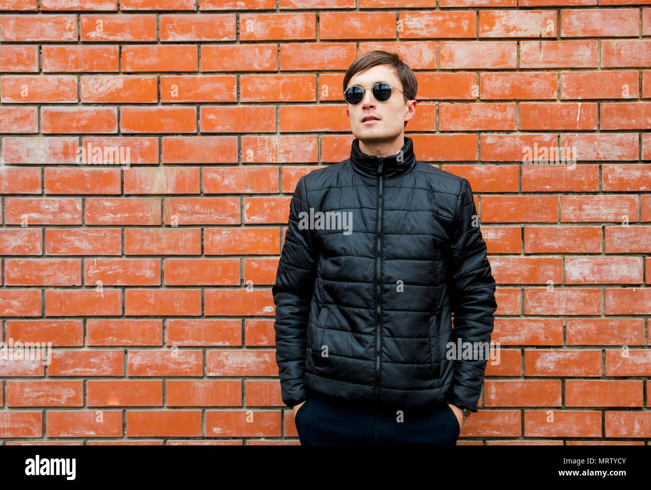 a young man stands leaning against a brick wall in sunglasses cross his ...