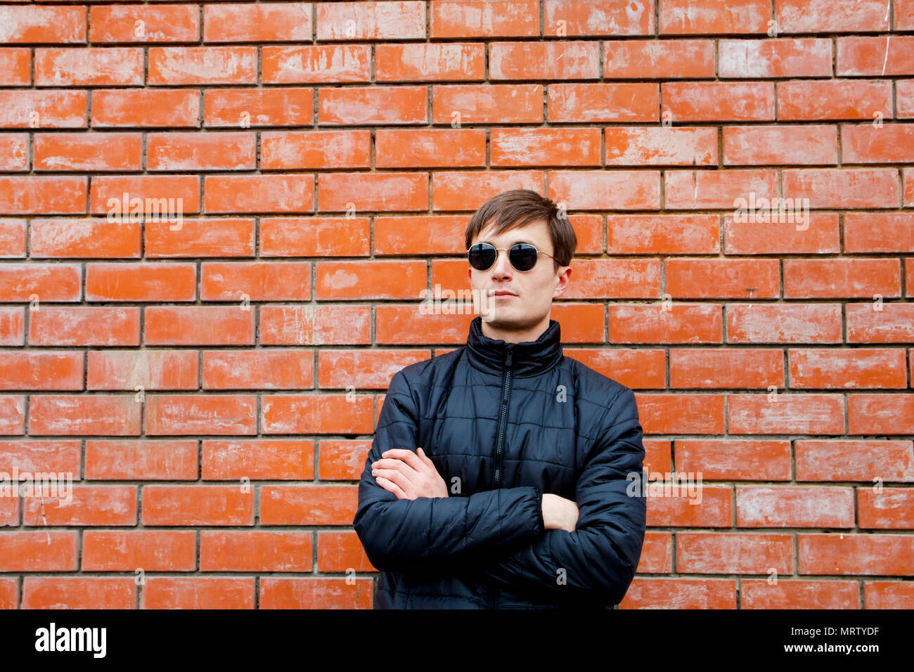 a young man stands leaning against a brick wall in sunglasses cross his arms. Stock Photo