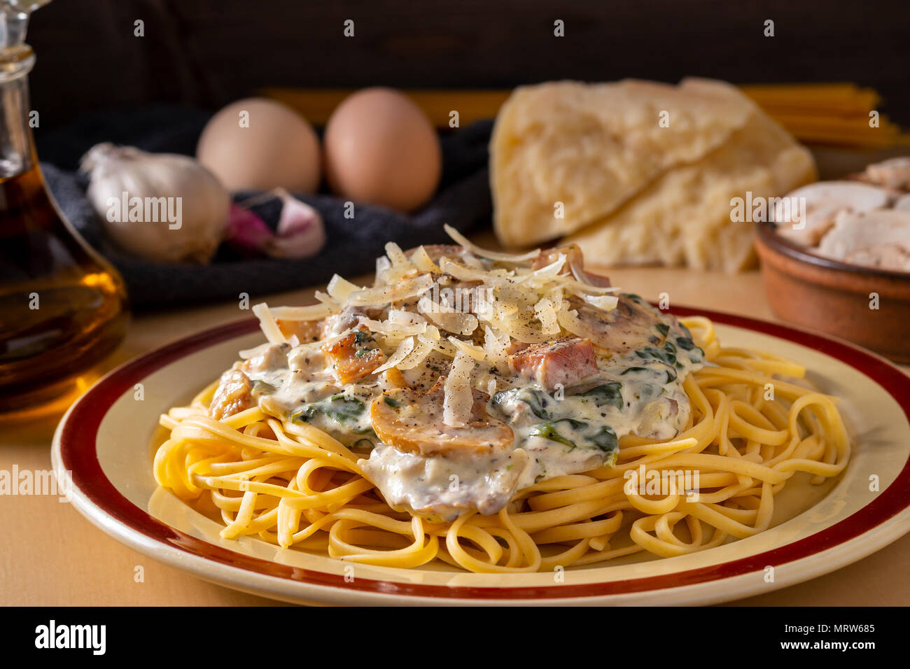 Plate with pasta topped with creamy sauce, beacon, spinach and mushrooms served on table with cooking ingredients Stock Photo