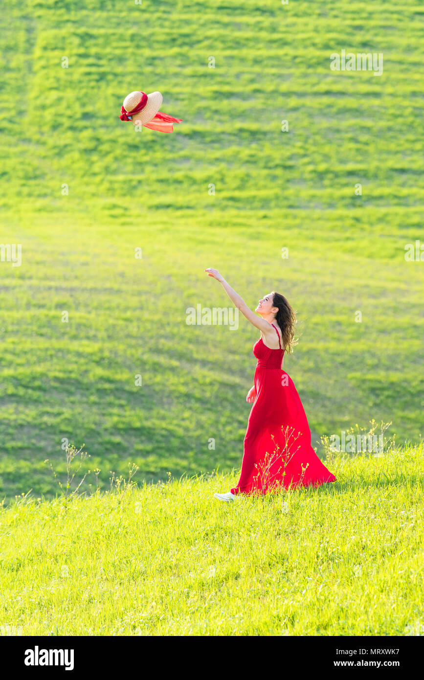 San Quirico d'Orcia, Orcia valley, Siena, Tuscany, Italy. A young woman in red dress  is throwing her hat in a wheat field Stock Photo