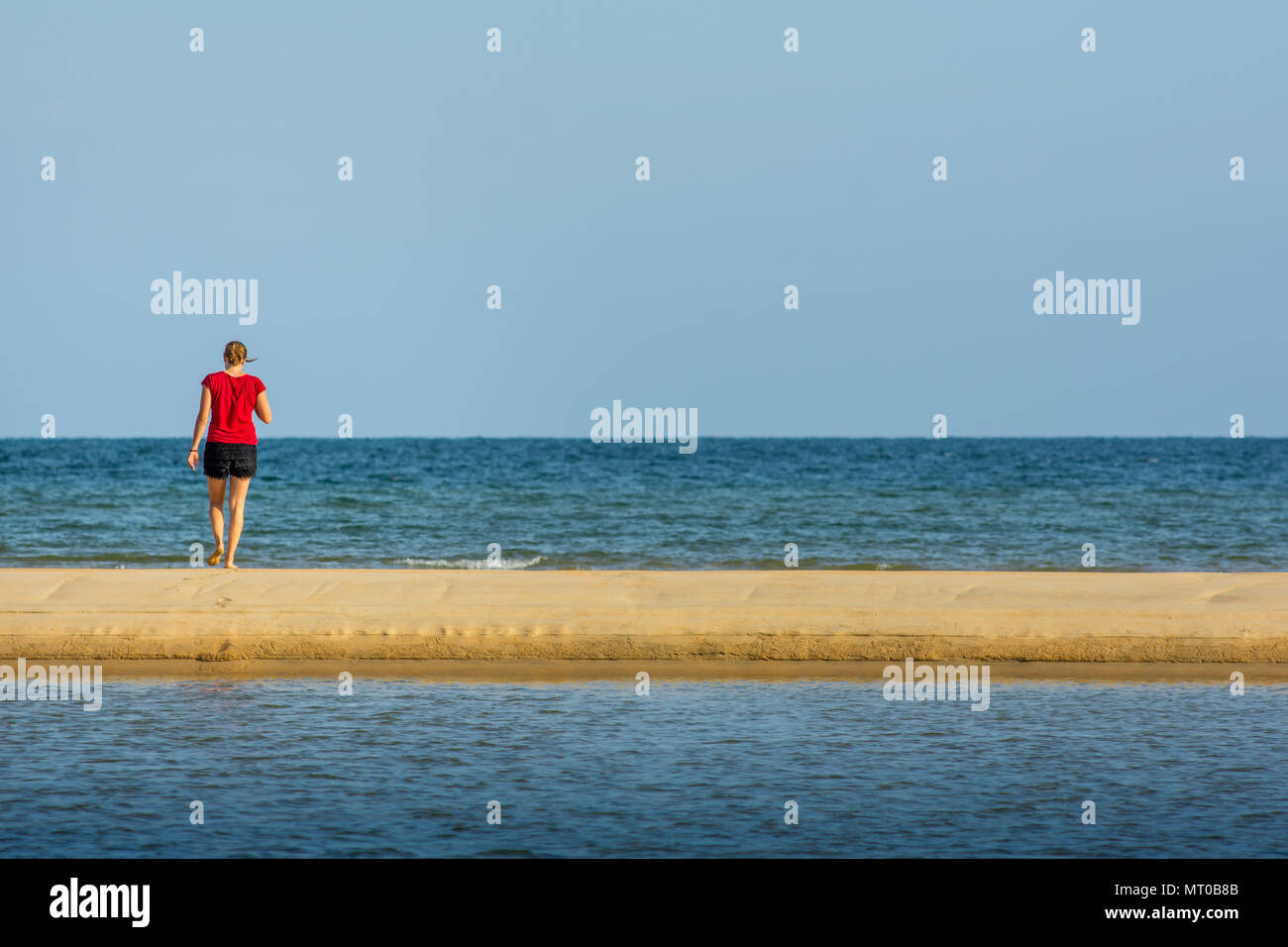 A young women walks along a sand back towards a calm sea. Stock Photo