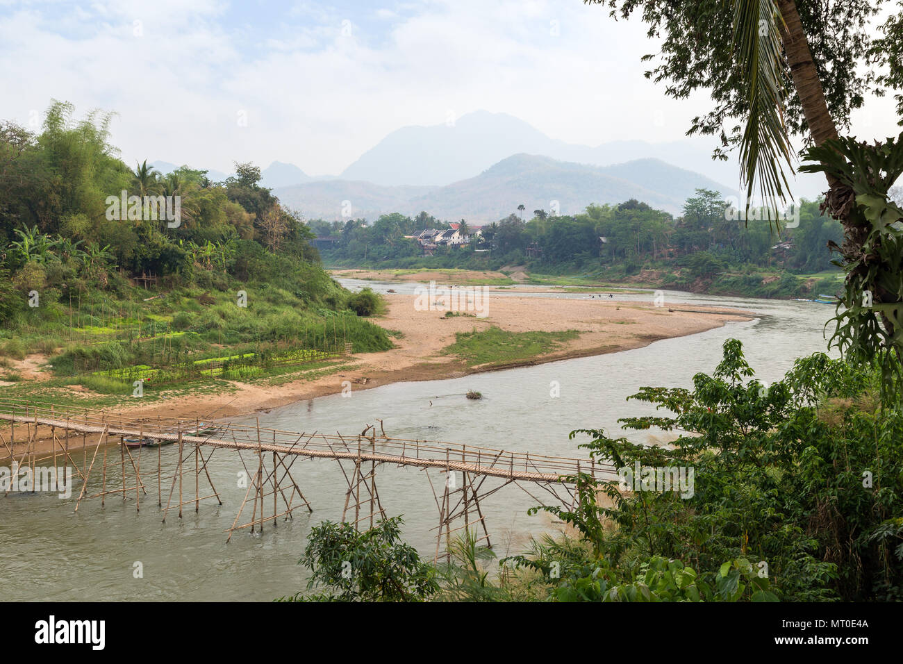View of a bamboo bridge over Nam Khan River at low tide and lush riverbank in Luang Prabang, Laos, on a sunny day. Stock Photo