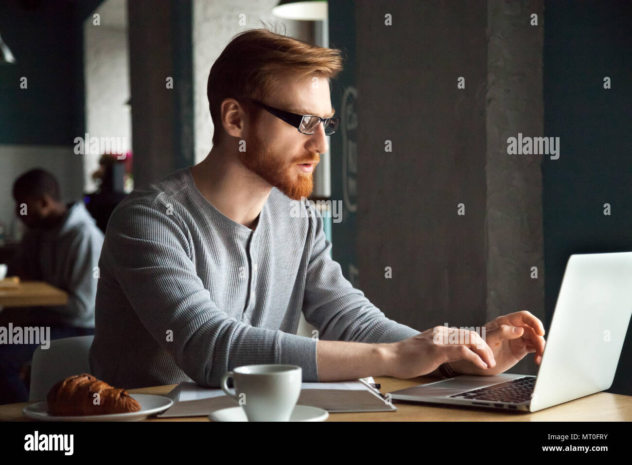 Focused millennial redhead man using laptop sitting at cafe tabl Stock Photo