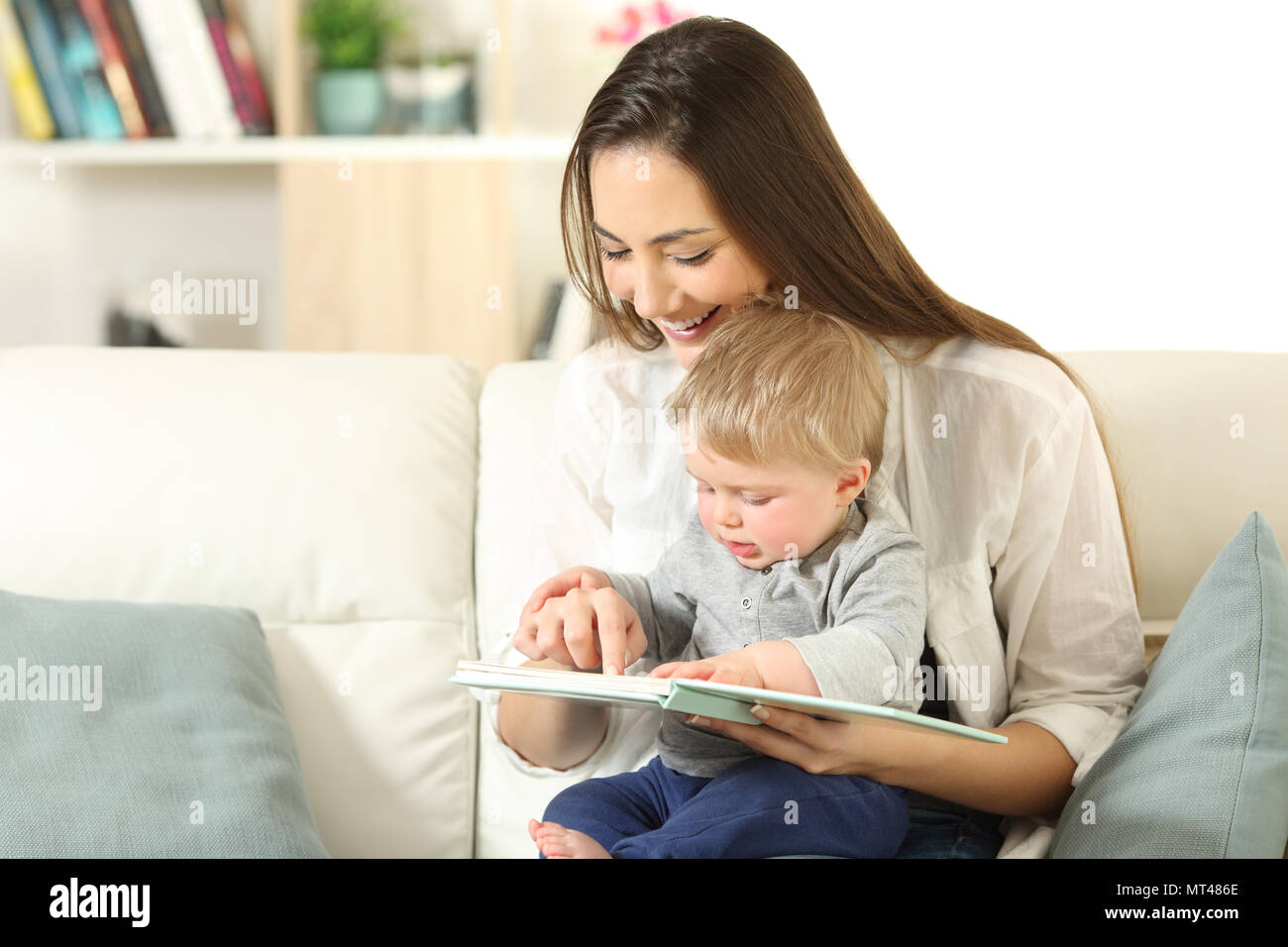 Baby and happy mother reading a book together sitting on a couch in the living room at home Stock Photo