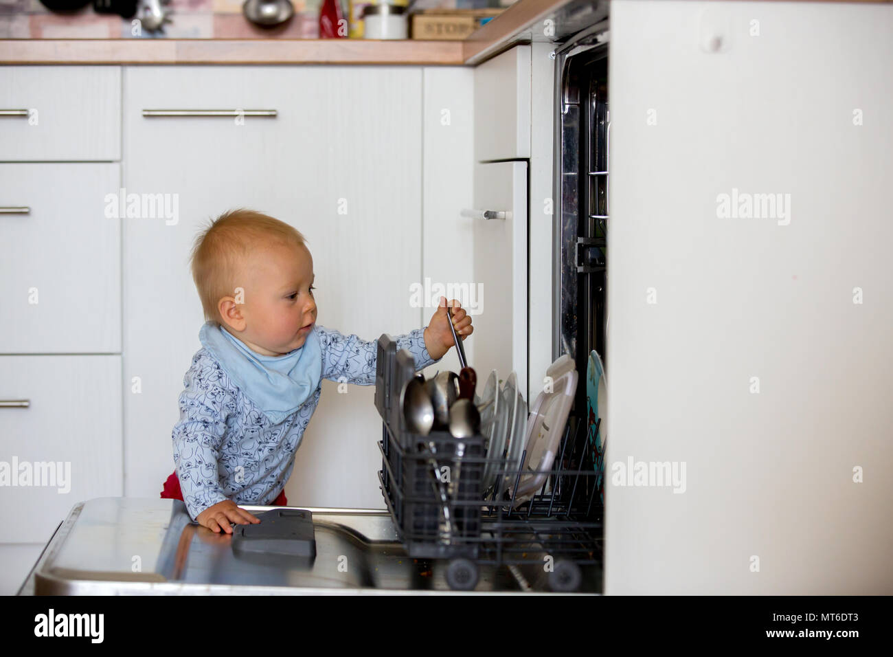 Toddler child, boy, helping mom, putting dirty dishes in dishwasher at home, modern kitchen Stock Photo