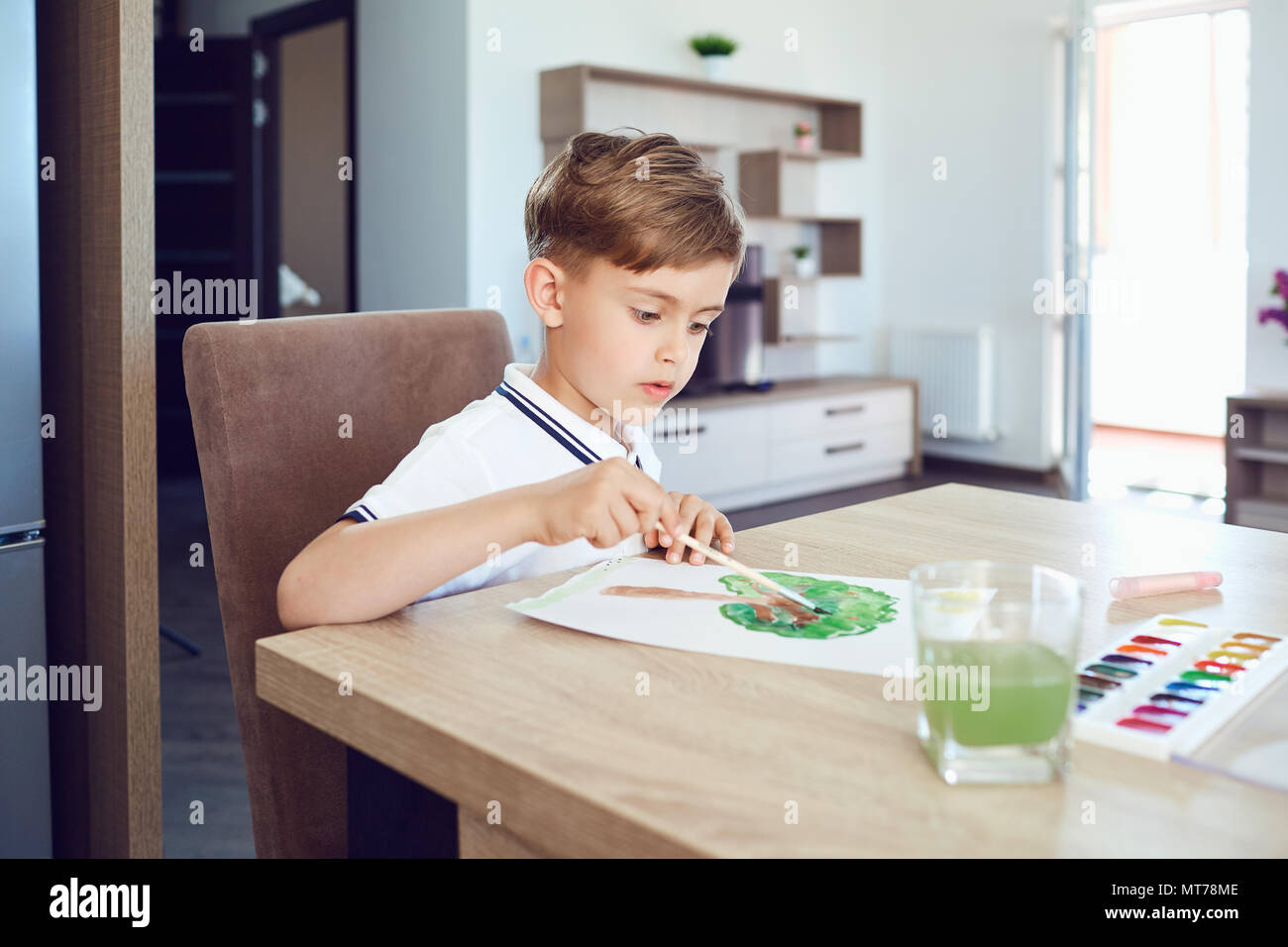 A blond boy draws on paper colored paints in the room. Stock Photo