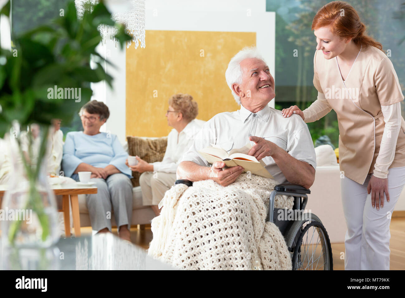 Happy paralyzed senior man in a wheelchair reading a book and a nurse helping him Stock Photo