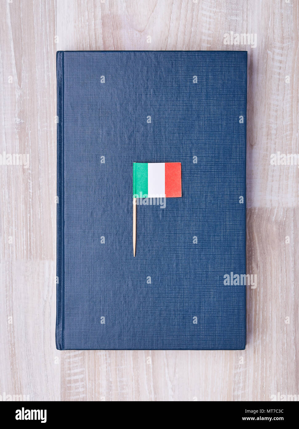 From above shot of simple blue notebook lying on wooden table with small Italian flag on toothpick on top. Stock Photo