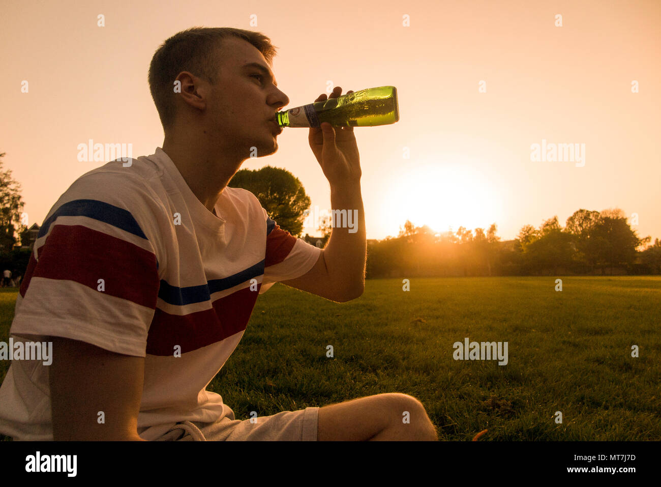 A teenager drinks from a beer bottle as the sun sets in an urban setting Stock Photo