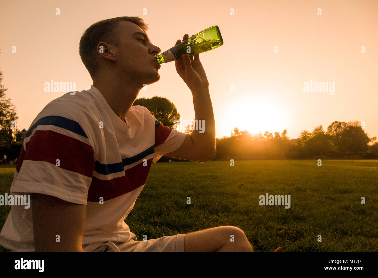 A teenager drinks from a beer bottle as the sun sets in an urban setting Stock Photo