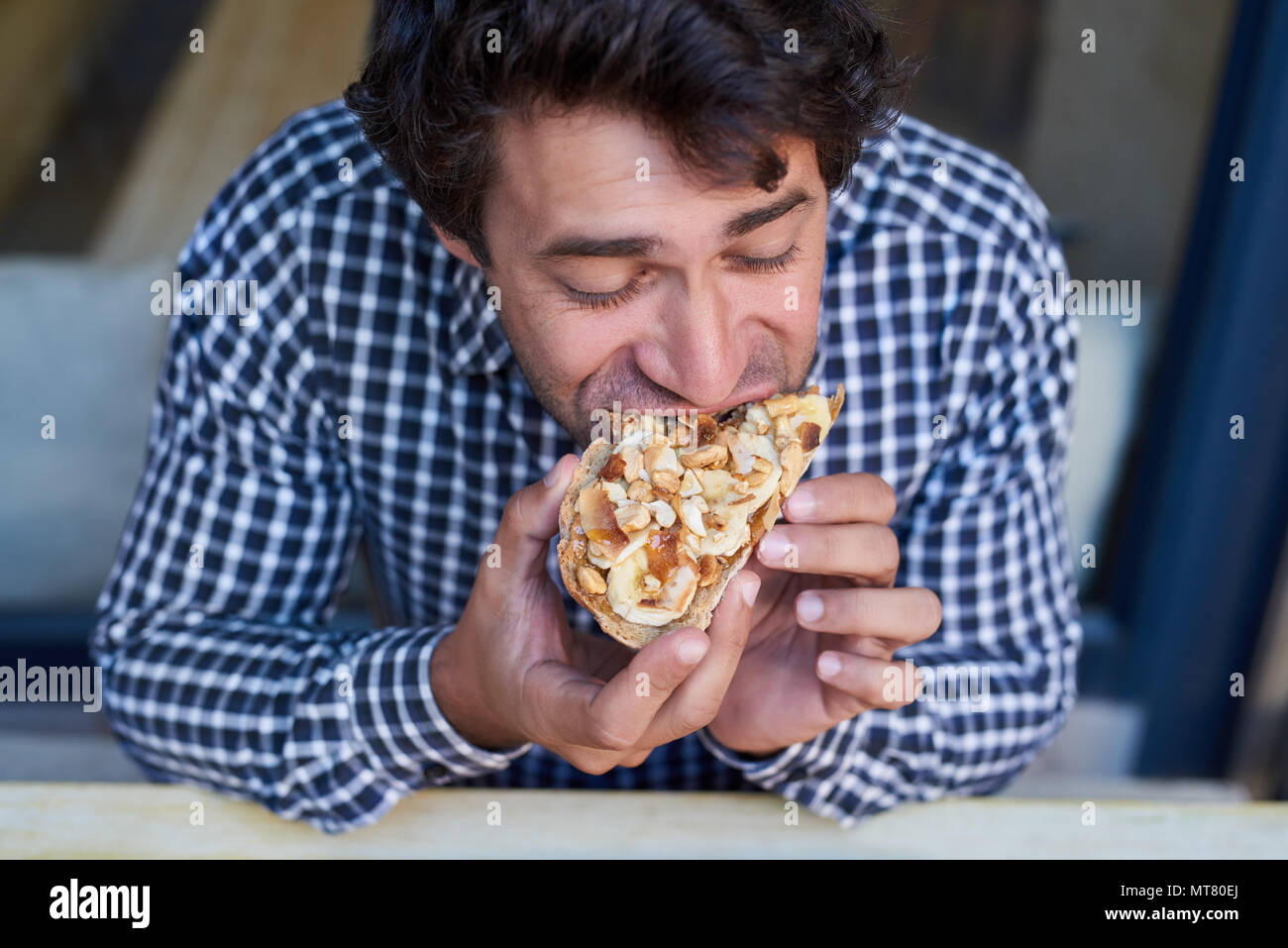 Hungry young man sitting at a bistro table taking a bite of his delicious peanut butter and banana open faced sandwich Stock Photo