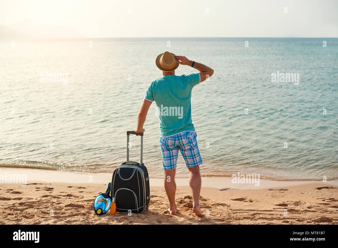Man tourist in summer clothes with a suitcase in his hand, looking at the sea on the beach, concept of time to travel Stock Photo