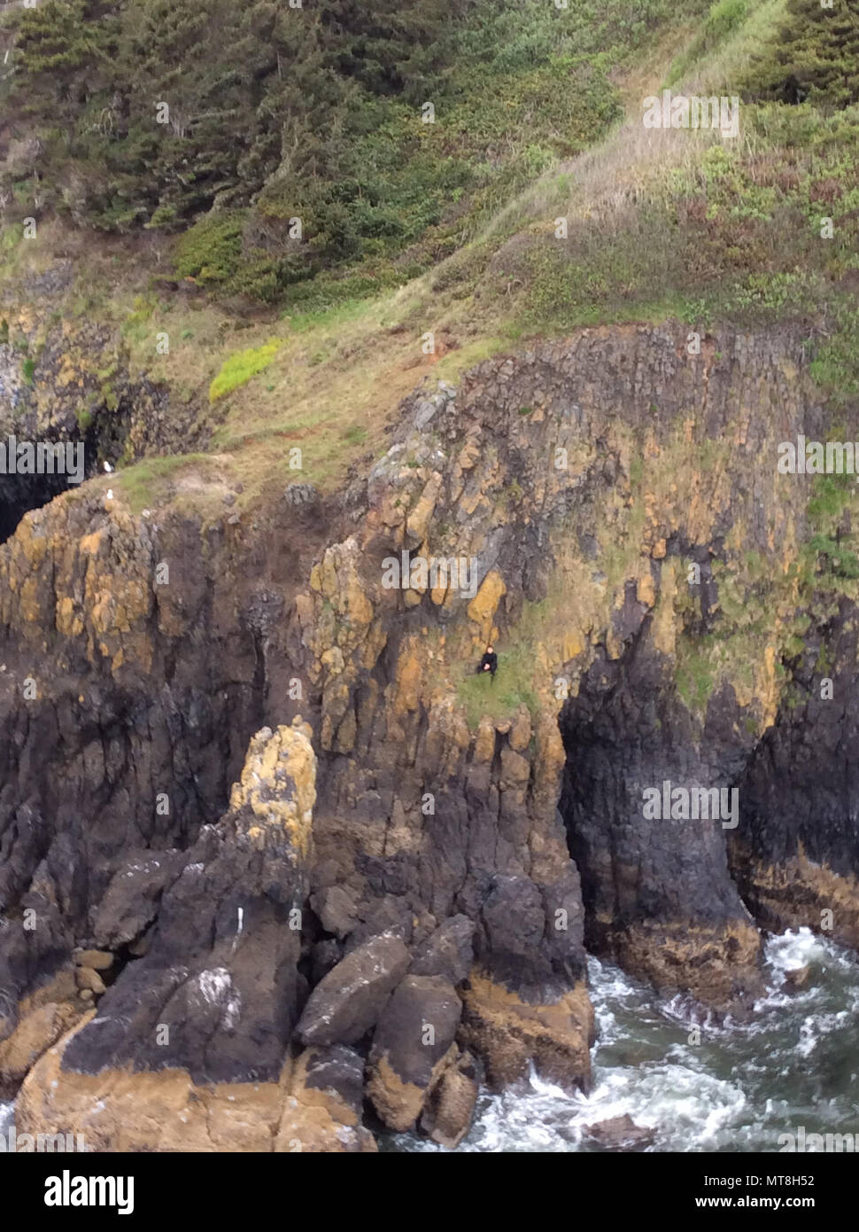 A surfer, in a black wetsuit, sits on a cliffside near Yaquina Head Lighthouse in Newport, Oregon, after he tried to climb to safety, May 13, 2018.    A Coast Guard aircrew aboard an MH-65 Dolphin helicopter form Air Facility Newport hoisted the man to safety and no major medical concerns were reported.    U.S. Coast Guard photo by Lt. Justin Bench. Stock Photo