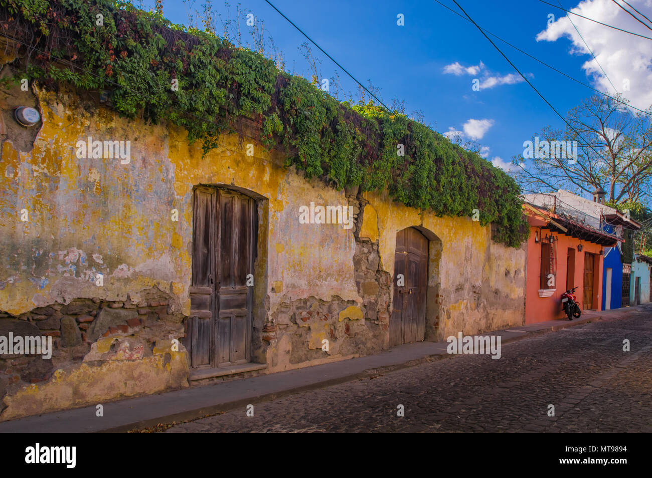 Outdoor view of yellow wall of old building, almost destroyed with some plants over the wall, close to colonial house buildings in Antigua city Stock Photo