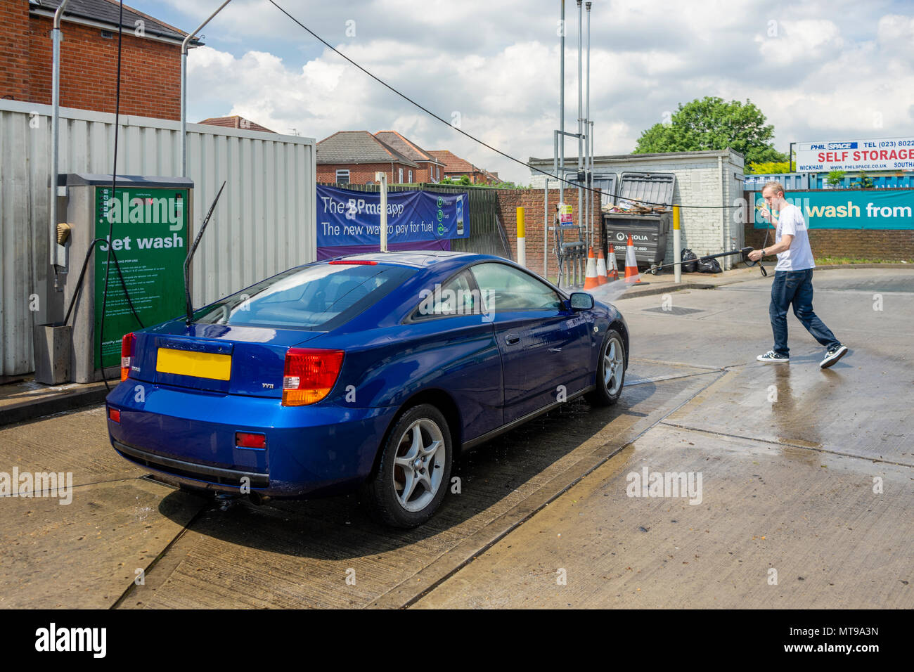 Male (age 30-40 years old) using a hand held jet wash pressure washer to clean a blue Toyata Celica car at a petrol station in the UK, Europe Stock Photo