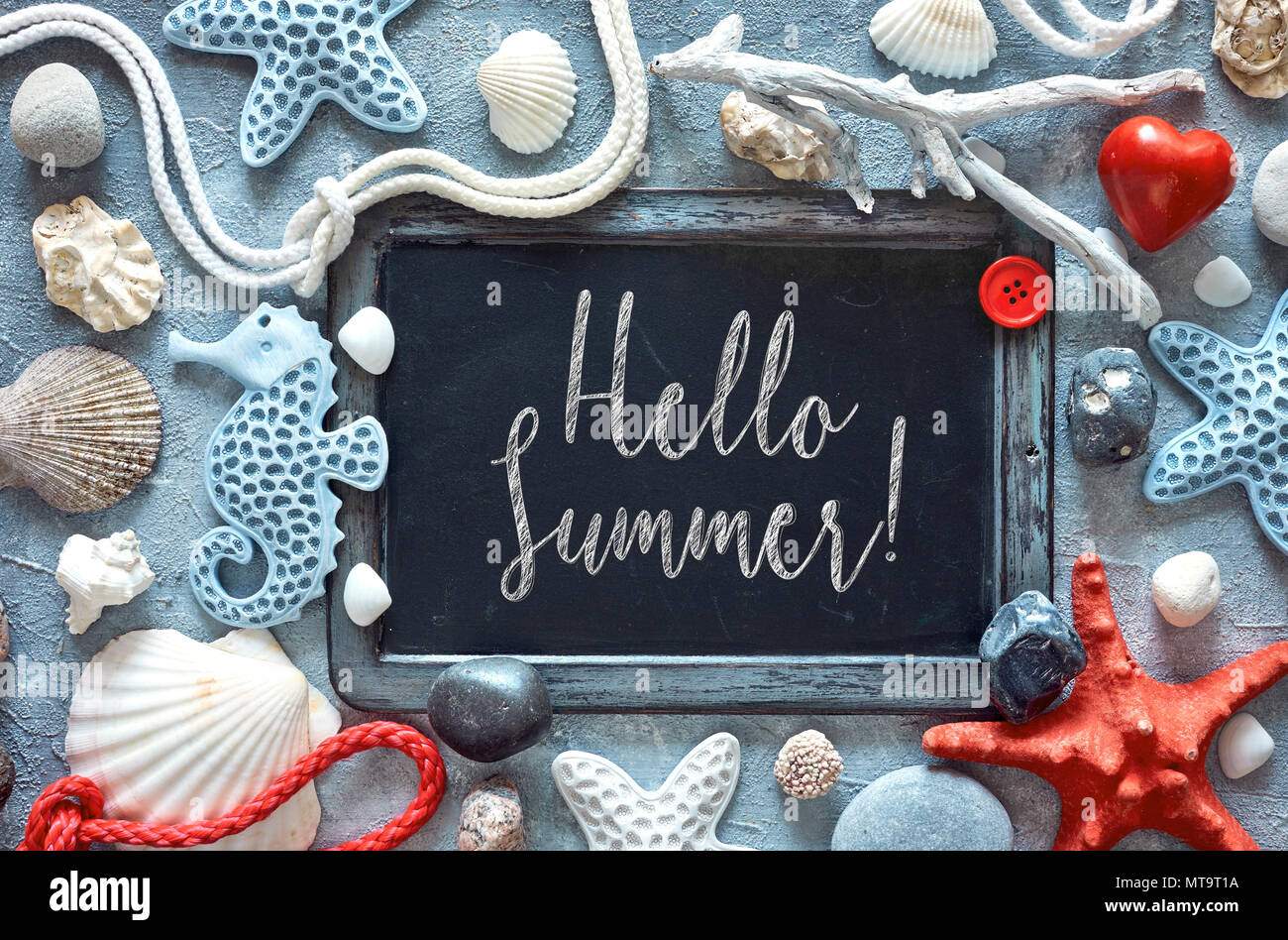 Blackboard with sea shells, stones, rope and star fish on textured light blue background, text 'Hello Summer' in chalk Stock Photo