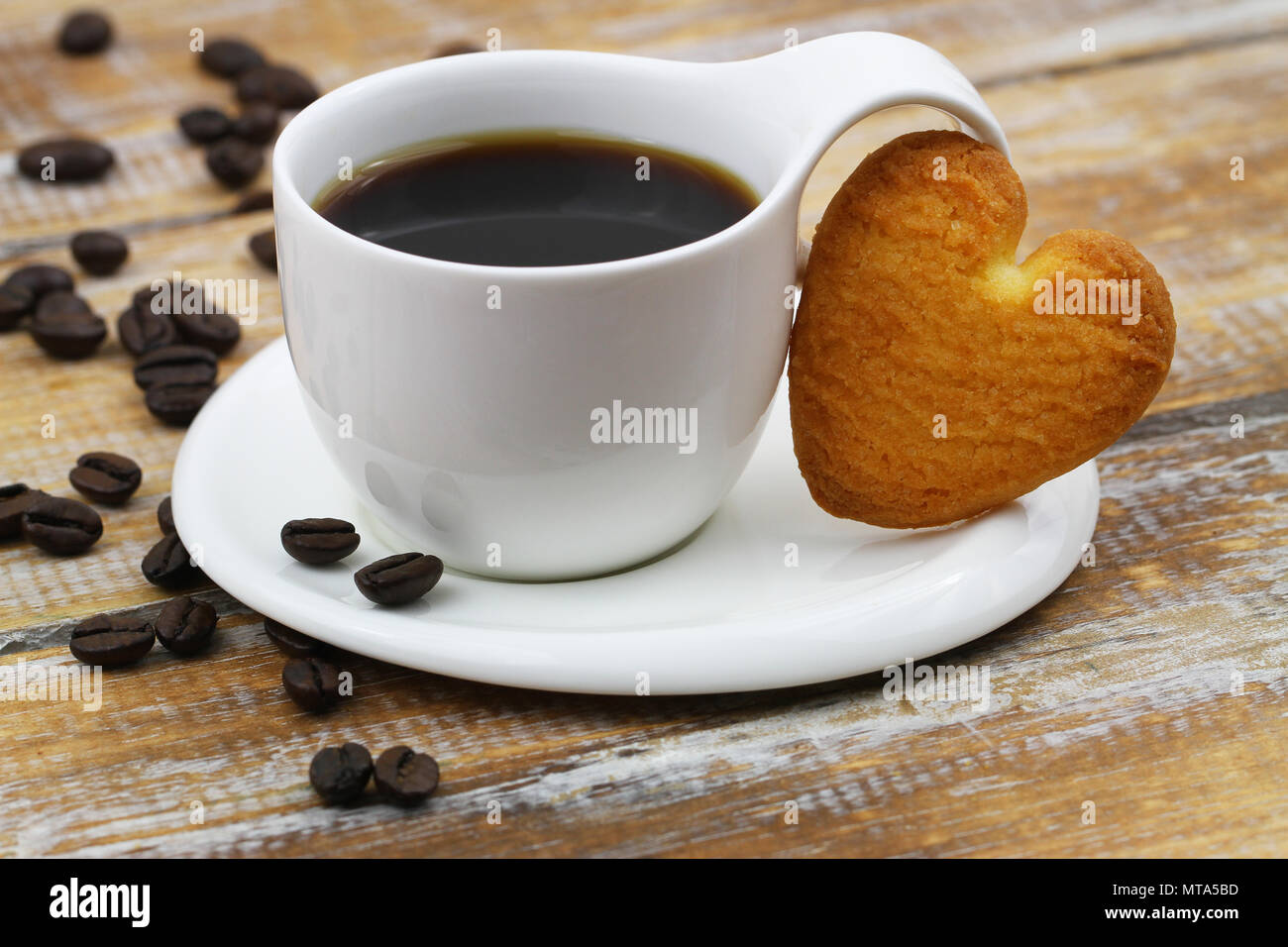 Heart shaped cookie leaning against white cup of coffee on rustic wooden surface Stock Photo