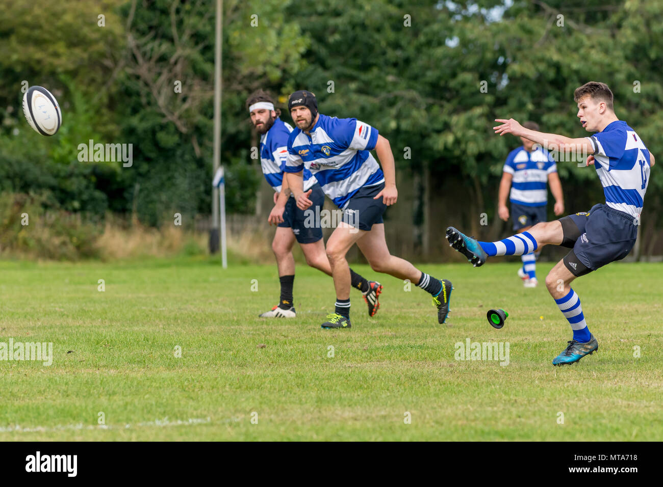 19 year old rugby player kicks ball for conversion, with teammates running forwards to chase the ball. The kicker, kicking tee and ball in shot. Stock Photo