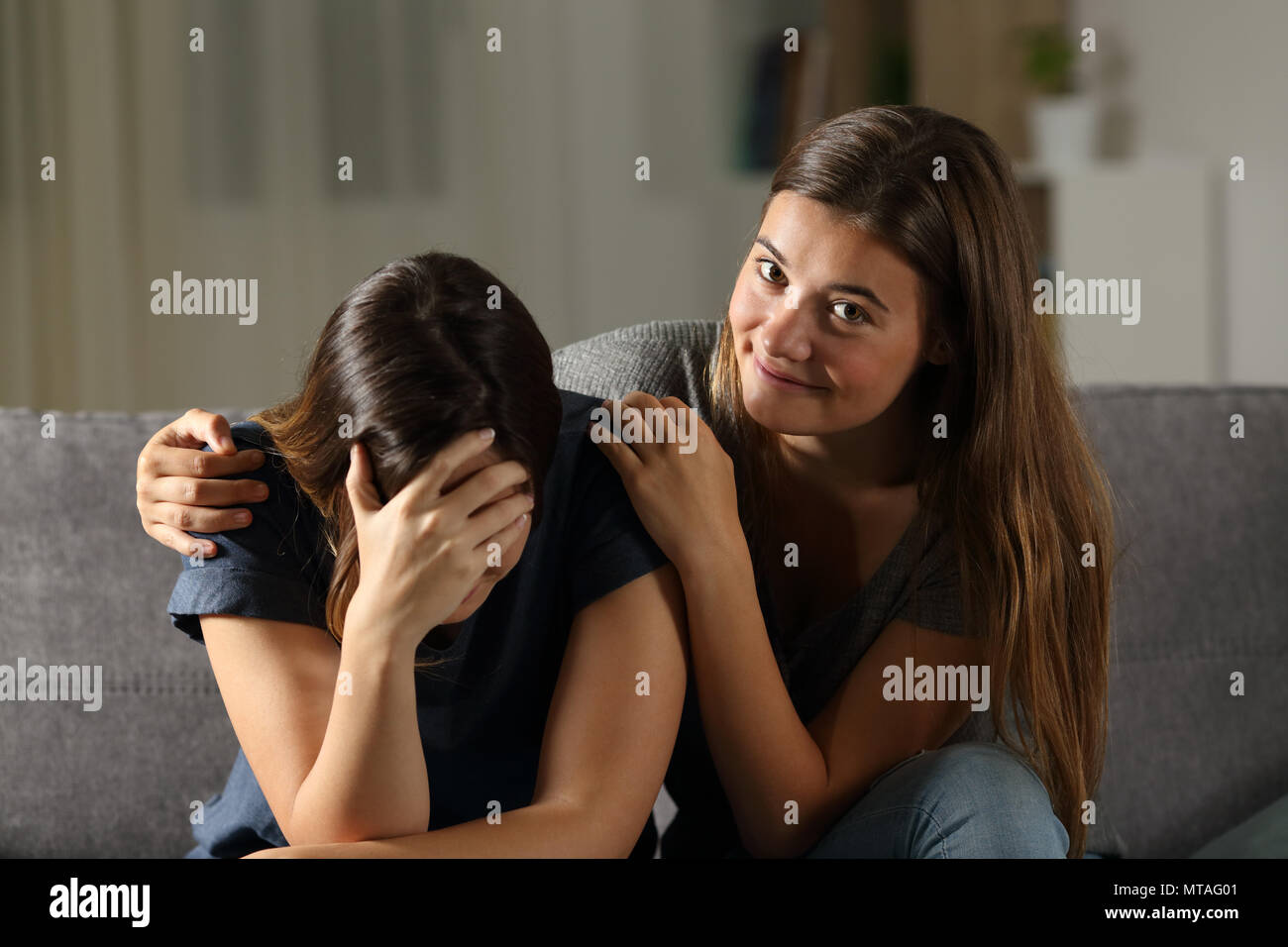 Bad teen is happy with her sad friend crying sitting on a couch in the living room at home Stock Photo