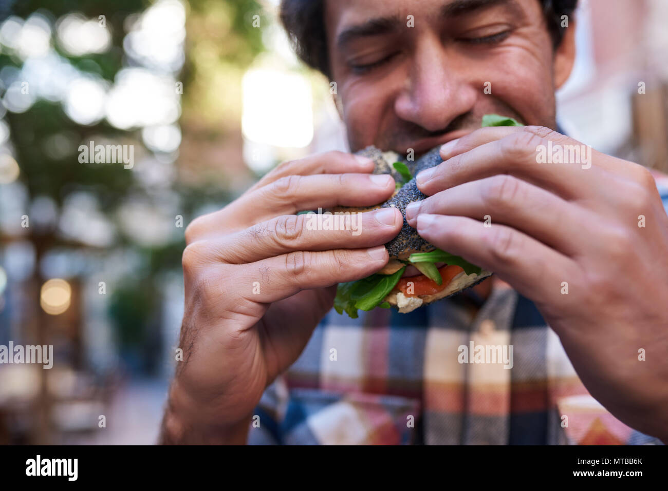 Closeup of a hungry young man taking a bite of his poppy seed bagel while standing with his eyes closed outside Stock Photo