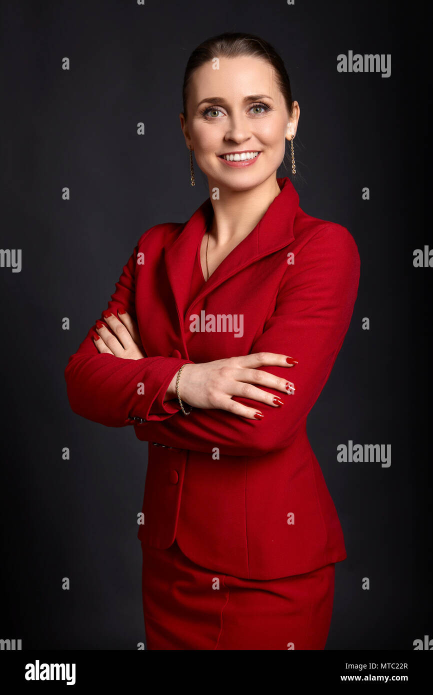 Portrait of elegant young business woman in red skirt suit with white smile and crossed arms, on black background. Stock Photo