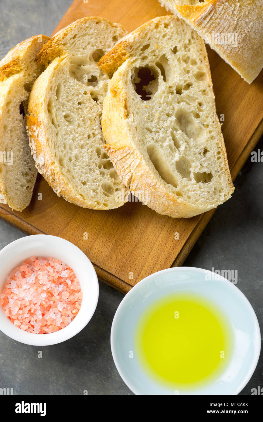 Top View of Sliced Artisan Rustic Bread on Wooden Cutting Board with Salt and Olive Oil on Dark Concrete Table. Golden Crust Appetizing Spongy Holey T Stock Photo