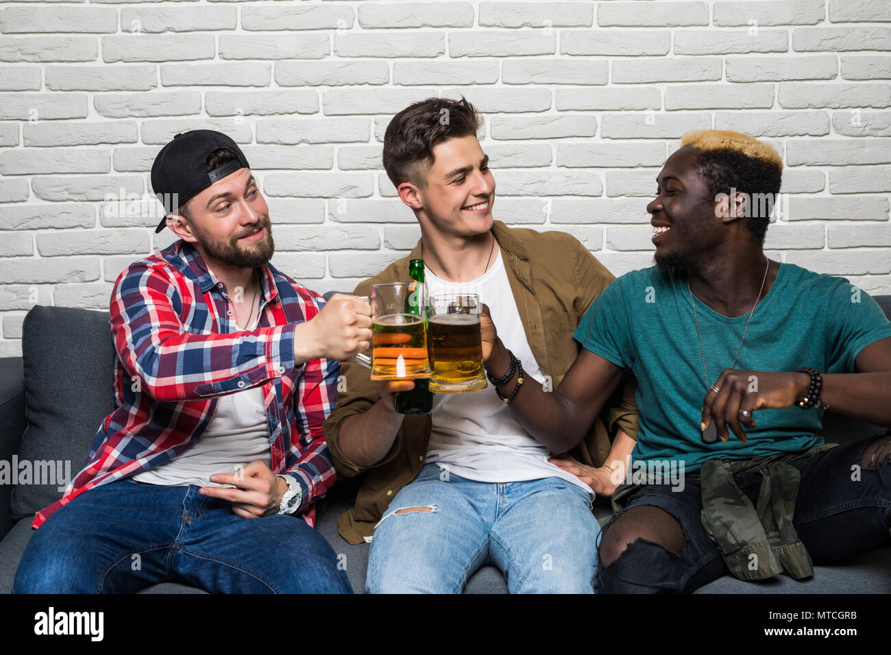 Domestic fans. Three happy young men watching football game cheers beer with pizza while sitting on sofa Stock Photo
