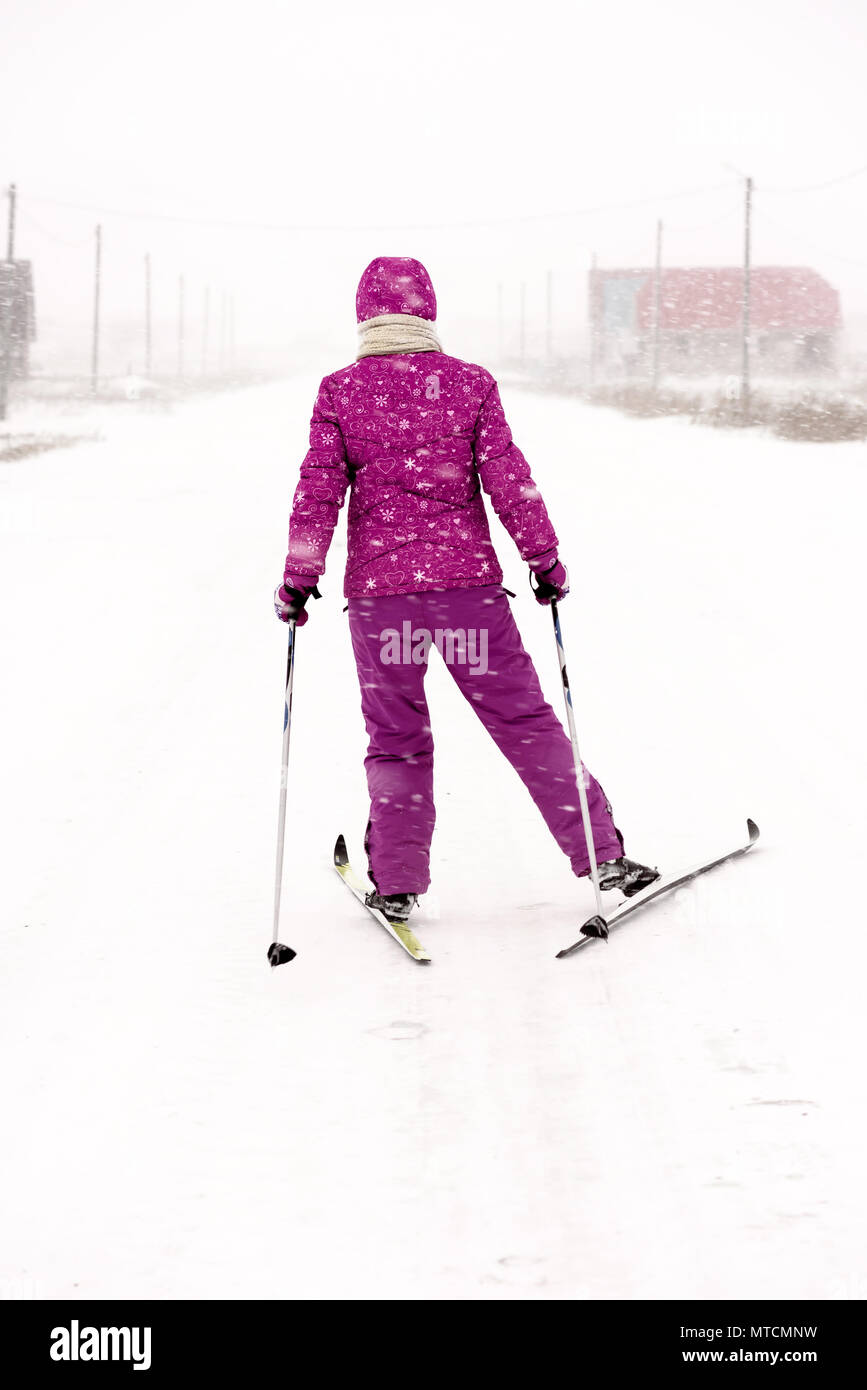 Young woman dressed in purple snow clothes and suit cross country skiing on a village road in a strong snow blizzard in a wintry snow swept scene and  Stock Photo