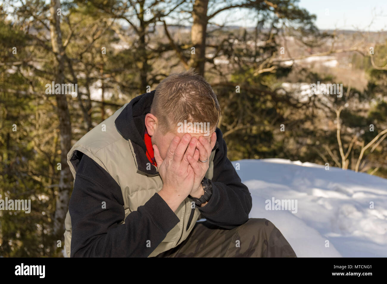 Mid adult 40s caucasian man sitting down outdoors in snow in winter with head in hands looking upset Stock Photo