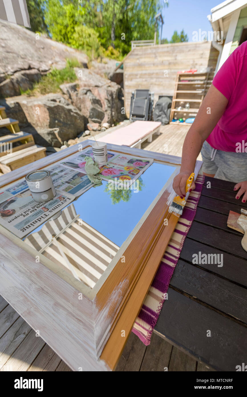 Adult woman outdoors on patio doing DIY and painting a wooden mirror frame with white paint Stock Photo