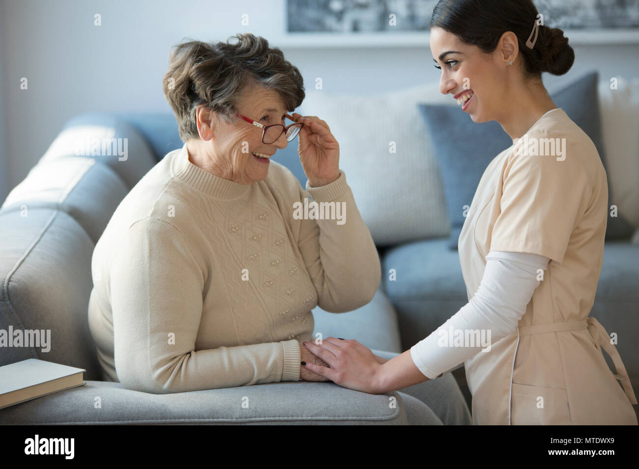 A tender caregiver kneeling down and facing a smiling older woman who is sitting on a sofa and holding her glasses Stock Photo