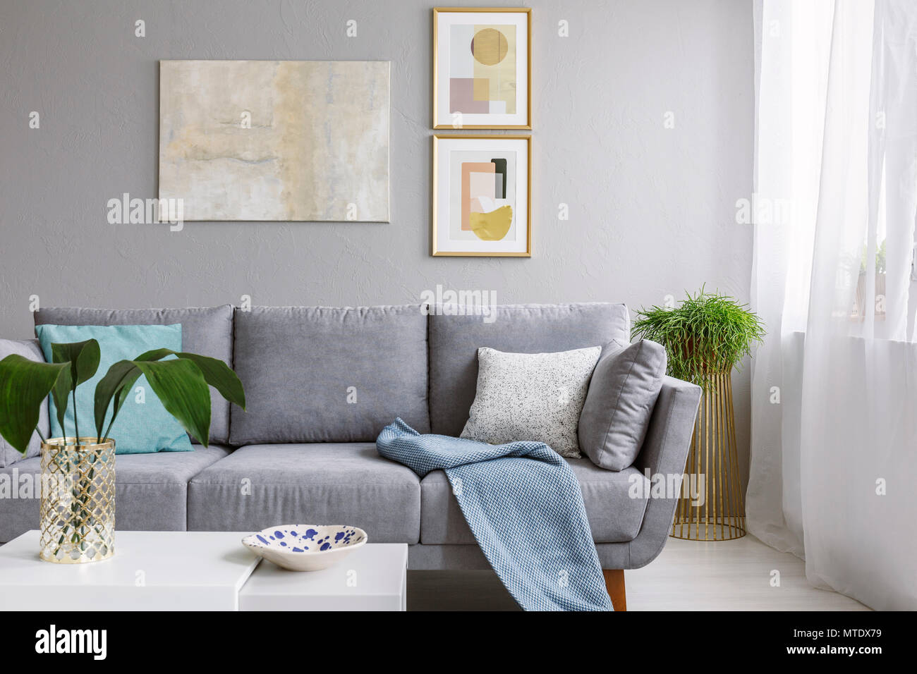Real photo of a grey sofa standing in a stylish living room interior behind a white table with leaves and in front of a grey wall with posters Stock Photo