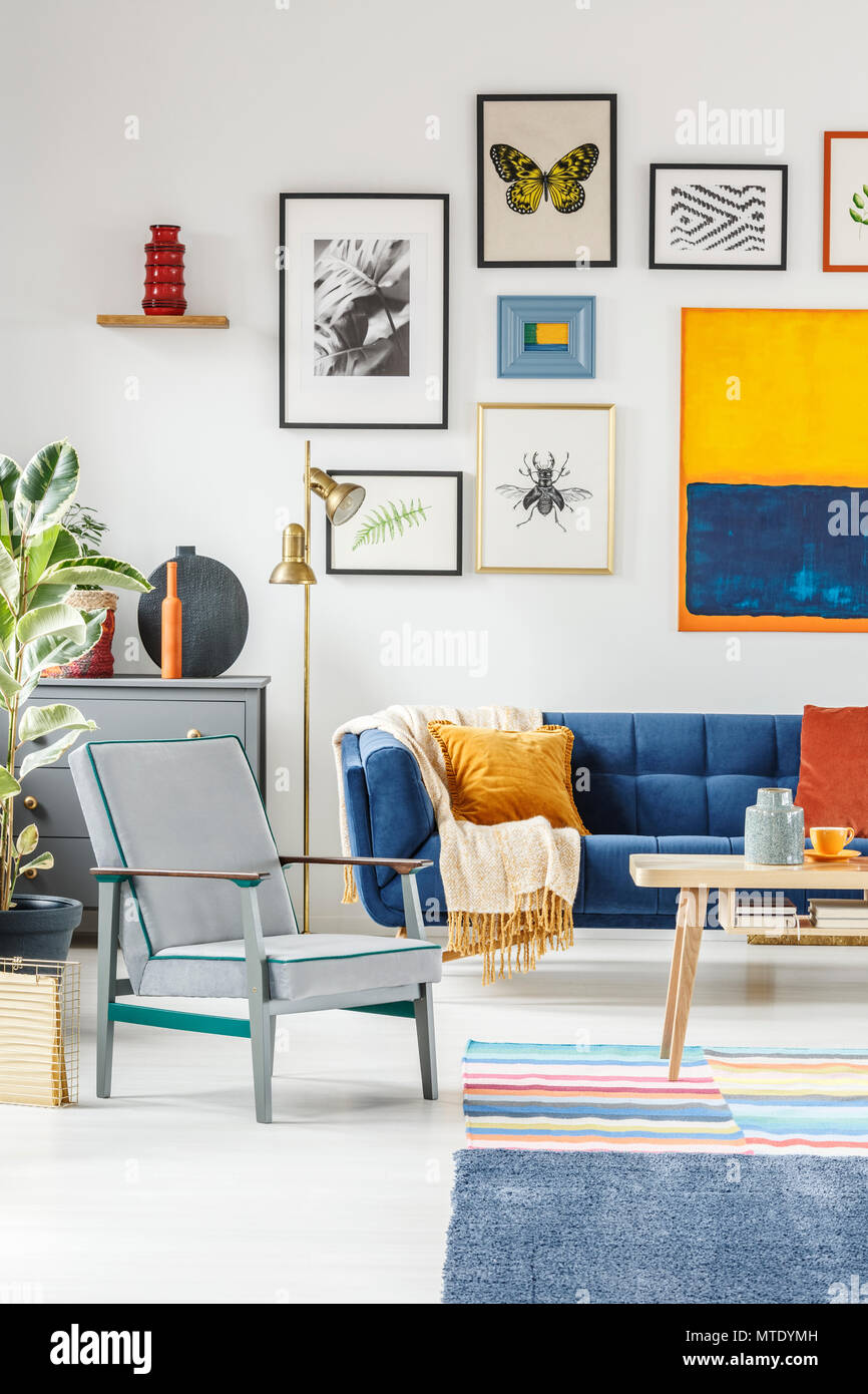 Real photo of a boho living room interior with a gallery of posters hanging on white wall above a gray armchair and blue couch Stock Photo